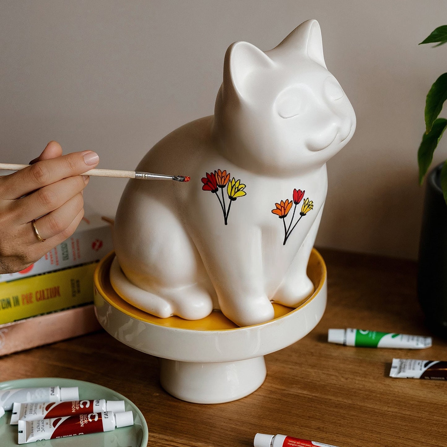 White ceramic cat urn sculpture on a wooden table with paint tubes and a hand holding a brush. Features personalized cat urn for ashes, custom cat cremation urn, cat-shaped urn, sleeping cat statue urn, angel cat urn, and pet urns for cats with engravable and personalized options.