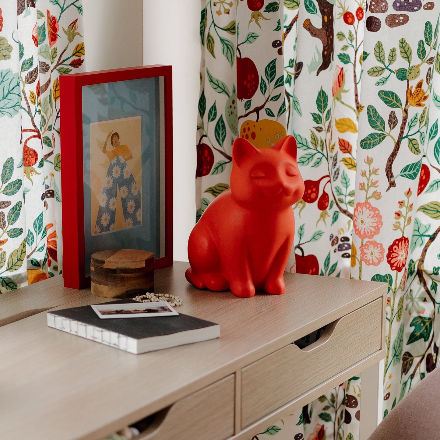 Red ceramic cat figurine cremation urn on a wooden surface with floral-patterned curtains in the background. Features personalized cat urn for ashes, custom cat cremation urn, cat-shaped urn, sleeping cat statue urn, and pet urns for cats with engravable and personalized options.