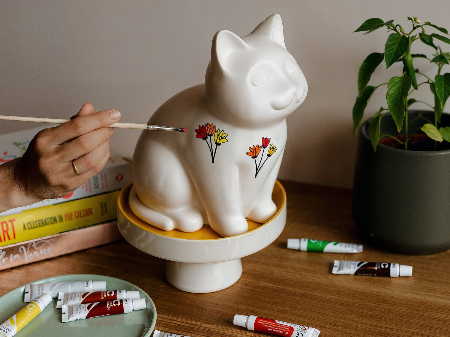 White ceramic cat urn sculpture on a wooden table with paint tubes and a hand holding a brush. Features personalized cat urn for ashes, custom cat cremation urn, cat-shaped urn, sleeping cat statue urn, angel cat urn, and pet urns for cats with engravable and personalized options.