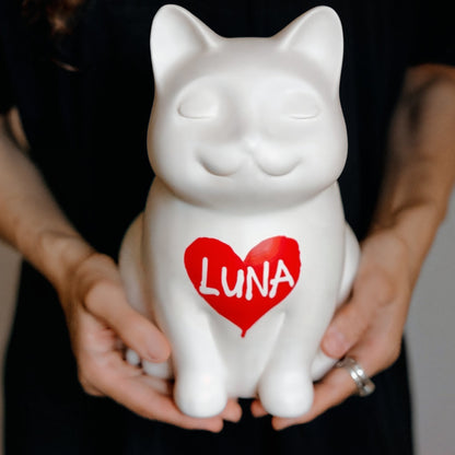 White cat-shaped ceramic urn container with a red heart and 'LUNA' text, held by hands against a black background. Features personalized cat urn for ashes, custom cat cremation urn, cat-shaped urn, angel cat urn, sleeping cat statue urn, and pet urns for cats with engravable and personalized options.