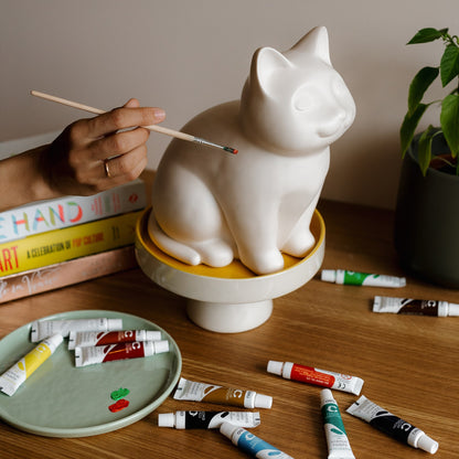 White ceramic cat urn sculpture on a wooden table with paint tubes and a hand holding a brush. Features personalized cat urn for ashes, custom cat cremation urn, cat-shaped urn, sleeping cat statue urn, angel cat urn, and pet urns for cats with engravable and personalized options.
