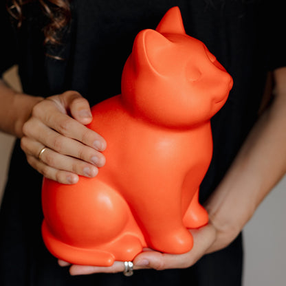 Orange cat-shaped ceramic cremation urn held by a person against a dark background. Features personalized cat urn for ashes, custom cat cremation urn, cat-shaped urn, sleeping cat statue urn, and pet urns for cats with engravable and personalized options.