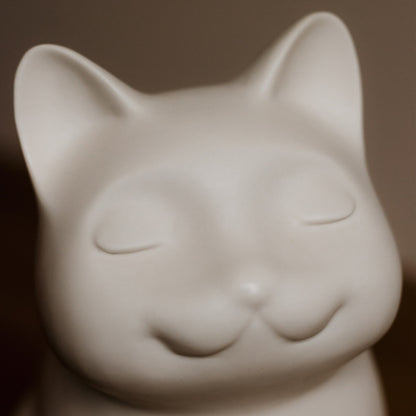 Close-up of a white ceramic cat-shaped urn on a blurred background. Features personalized cat urn for ashes, custom cat cremation urn, cat-shaped urn, sleeping cat statue urn, and pet urns for cats with engravable and personalized options.