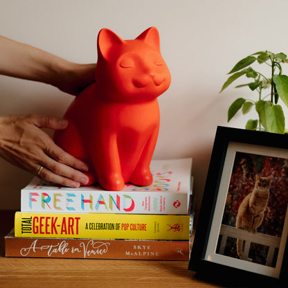 Red cat-shaped ceramic urn for ashes on a stack of books with a framed picture of a cat in the background. Features personalized cat urn for ashes, custom cat cremation urn, cat-shaped urn, sleeping cat statue urn, and pet urns for cats with engravable and personalized options.
