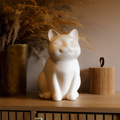 White ceramic cat urn on a wooden surface with decorative plants in the background. Features personalized cat urn for ashes, custom cat cremation urn, cat-shaped urn, sleeping cat statue urn, and pet urns for cats with engravable and personalized options.