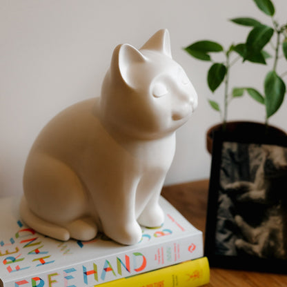 White ceramic cat urn on a stack of books with a plant in the background. Features personalized cat urn for ashes, custom cat cremation urn, cat-shaped urn, sleeping cat statue urn, and pet urns for cats with engravable and personalized options.