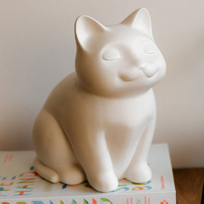 White ceramic cat figurine urn placed on a colorful book. Features personalized cat urn for ashes, custom cat cremation urn, cat-shaped urn, sleeping cat statue urn, and pet urns for cats with engravable and personalized options.